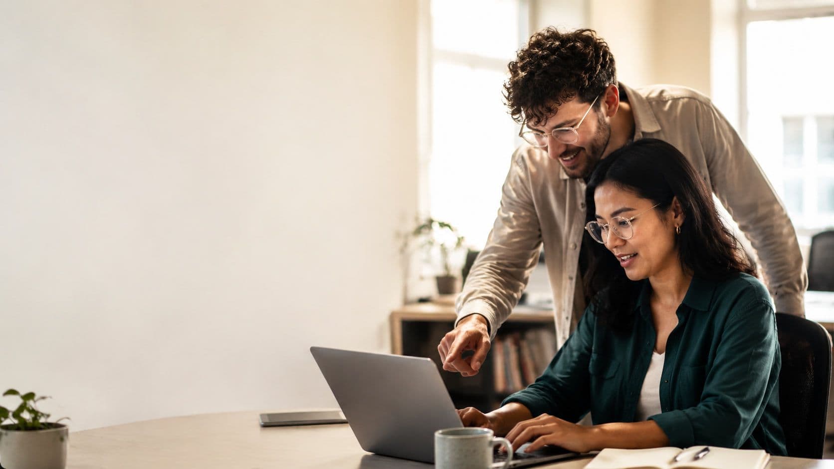 Two colleagues at a laptop in a bright office, one leaning in to point at the screen while the other types